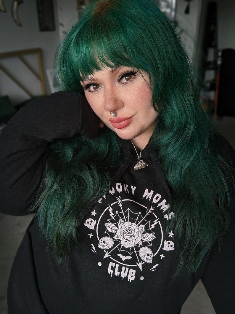 以下が含まれることがあります： 白いグラフィックプリントのバラ、頭蓋骨、そして「Spooky Moms Club」というテキストが書かれた黒いパーカー。モデルは緑色の髪をしています。