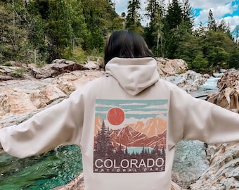 Puede incluir: Una sudadera con capucha beige con un gráfico de estilo vintage de una cordillera y un sol rojo. El gráfico incluye el texto "Colorado National Park".