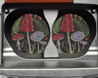 May include: Two round black car coasters with a mushroom and fern design. The mushrooms are red with white spots and the ferns are green. The coasters are in a car cup holder.