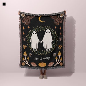 May include: Black woven throw blanket with a spooky design featuring two ghosts holding hands in a wreath of leaves. The blanket has a fringe border and the text "AVA & NATE" in the center.