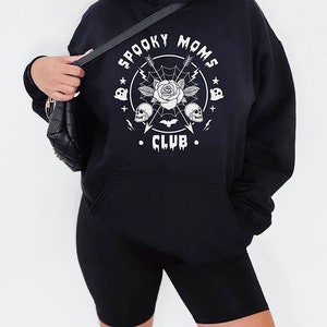 以下が含まれることがあります： 「Spooky Moms Club」という白いグラフィックプリントが施された黒いパーカー。バラ、ドクロ、コウモリ、矢印がデザインされています。