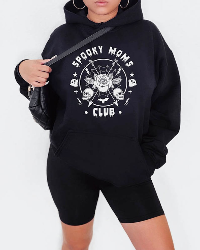 以下が含まれることがあります： 「Spooky Moms Club」という白いグラフィックプリントが施された黒いパーカー。バラ、ドクロ、コウモリ、矢印がデザインされています。