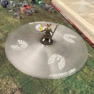 May include: A gaming setup for a tabletop role-playing game. A miniature figure stands on a gray circular mat with the text "SPIRIT GUARDIANS" and a repeating design. Dice and an open book are also visible, suggesting a fantasy game.