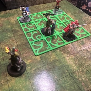 May include: A tabletop role-playing game setup featuring miniature figurines on a green grid. The figurines depict various characters, including a warrior with a red cape and a creature with horns. The game board has a green floral design.