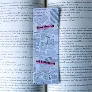 May include: Bookmark with a collage of newspaper clippings. The words "fund libraries" and "not billionaires" are written on pink tape across the bookmark. The bookmark is resting on an open book.