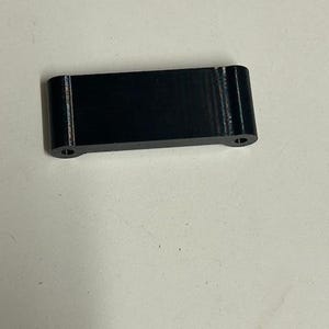 May include: A small, rectangular black object with rounded corners and two small holes on the ends. The object appears to be made of a solid material and has a matte finish. The item is likely a component or part.