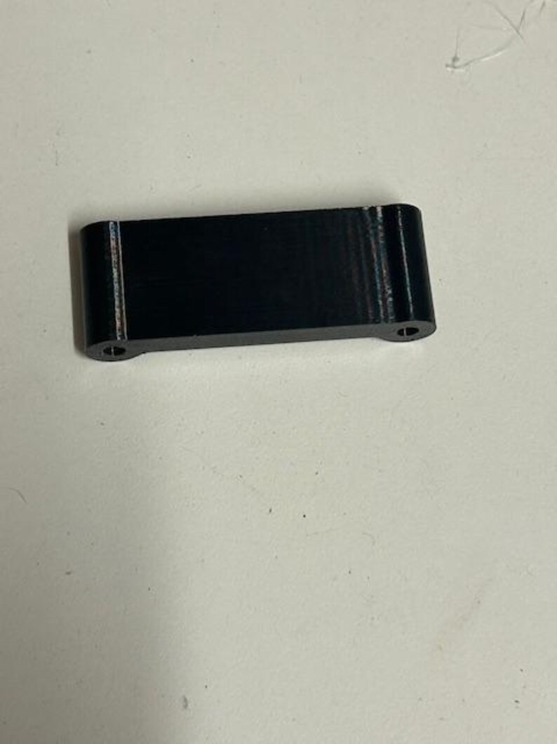 May include: A small, rectangular black object with rounded corners and two small holes on the ends. The object appears to be made of a solid material and has a matte finish. The item is likely a component or part.