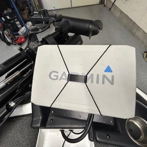 May include: A white Garmin fish finder with the brand name in gray lettering and a blue triangle logo. The device is secured with black bungee cords and mounted on a black surface, likely a boat console. A cup holder is visible.
