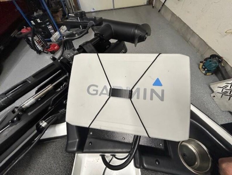 May include: A white Garmin fish finder with the brand name in gray lettering and a blue triangle logo. The device is secured with black bungee cords and mounted on a black surface, likely a boat console. A cup holder is visible.