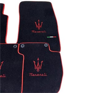 May include: Black car floor mats with red trim and the Maserati logo embroidered in red. The mats have a trident emblem and the Maserati name. An Italian flag is also present.