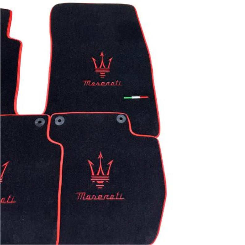 May include: Black car floor mats with red trim and the Maserati logo embroidered in red. The mats have a trident emblem and the Maserati name. An Italian flag is also present.