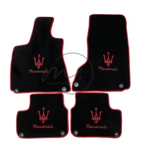May include: Set of four black car floor mats with red trim. Each mat features the Maserati logo in red. The mats are designed to fit a vehicle's front and rear footwells. The mats have a textured surface.