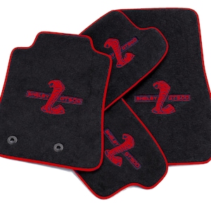 May include: Set of four black car floor mats with red trim. Each mat features a red cobra logo with "SHELBY GT500" embroidered in red. Two mats have mounting holes.