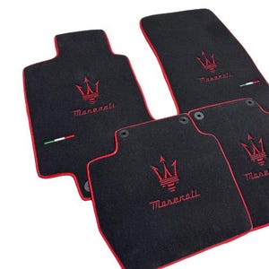 May include: Set of four black car floor mats with red trim. Each mat features the Maserati logo embroidered in red. The mats have an Italian flag detail and are designed to protect the vehicle's interior.
