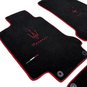 May include: Black car floor mats with red trim and the Maserati logo embroidered in red. The mats have a textured surface and include the Italian flag colors. The mats are designed to fit a car.