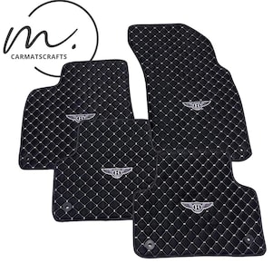 May include: Black car floor mats with a diamond-stitched pattern and white stitching. Each mat features a silver emblem. The logo "CARMATSCRAFTS" is in the upper left corner.