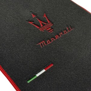 May include: Black car mat with a red border. Features the Maserati trident logo and the brand name embroidered in red. Includes a small embroidered Italian flag.