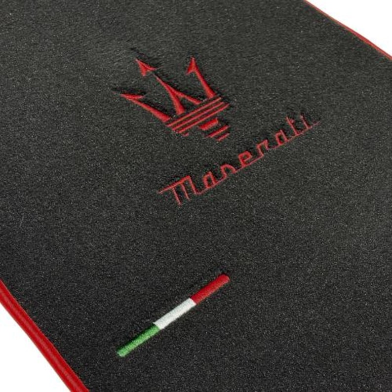 May include: Black car mat with a red border. Features the Maserati trident logo and the brand name embroidered in red. Includes a small embroidered Italian flag.