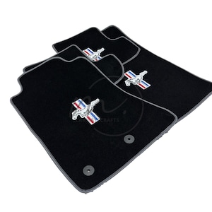 May include: Black car floor mats with a gray border. Each mat features a silver emblem of a running horse with red, white, and blue stripes. The mats are designed to fit a vehicle's interior and protect the floor.