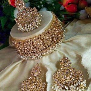Könnte beinhalten: Ein goldfarbenes Brautschmuck-Set mit Halskette, Ohrringen und Kopfschmuck. Das Set ist mit klaren Steinen und kleinen weißen Perlen verziert, wodurch ein elegantes Design entsteht. Der Schmuck wird auf einer cremefarbenen Oberfläche präsentiert.