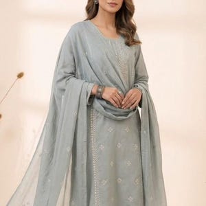 May include: A light sage green three-piece outfit consisting of a long-sleeved straight-cut tunic, matching straight-leg trousers, and a sheer dupatta. The tunic and dupatta feature delicate white embroidered patterns and subtle silver detailing.