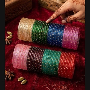 May include: A collection of colourful glass bangles in gold, green, blue, and pink, decorated with tiny gold dots. The bangles are displayed on a deep red surface, with spices scattered around.
