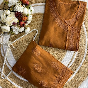 Può includere: Un completo a due pezzi in una calda tonalità terrosa. La parte superiore presenta ricami intricati intorno allo scollo e sul davanti. Anche i pantaloni abbinati hanno ricami floreali. Il set è esposto su un tappeto intrecciato con un bouquet di fiori e una collana di perle.