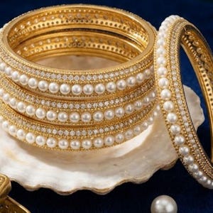 May include: Gold bangles with pearl and rhinestone accents. The stack of bangles is displayed on a white seashell against a dark blue background. The bangles have a classic design, suitable for special occasions.