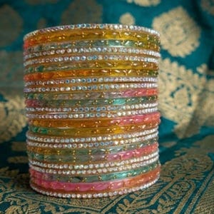 Puede incluir: Una pila de pulseras coloridas en tonos rosa, naranja, amarillo, verde y transparente. Cada pulsera está adornada con pequeños adornos brillantes. Las pulseras están apiladas sobre un fondo turquesa con un diseño floral dorado.