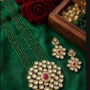 May include: A green beaded necklace and matching earrings set. The necklace features a large, round pendant with a central red stone surrounded by clear, geometric-shaped stones. The earrings have a similar design. The jewelry rests on a green fabric.