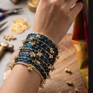 Könnte beinhalten: Ein Stapel dunkelblauer Armreifen mit goldenen Akzenten und kleinen goldenen Anhängern. Die Armreifen befinden sich an einem Handgelenk, mit einem goldenen Ring mit einem blauen Stein an einem Finger. Der Hintergrund umfasst Schmuckherstellungswerkzeuge und goldene Verzierungen.