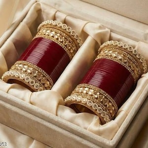 Op de afbeelding: Twee sets donkerrode armbanden met goudkleurige metalen banden, voorzien van bladvormige versieringen en rijen kleine witte parels. De armbanden zijn gerangschikt in een rechthoekige doos bekleed met crèmekleurige satijnen stof.