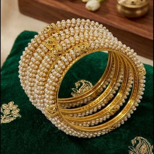 May include: A set of gold bangles with intricate detailing. The bangles are adorned with small, white pearls and ornate gold accents. The jewelry rests on a dark green velvet surface, creating a luxurious aesthetic.