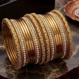 以下が含まれることがあります： 精巧なディテールと白い宝石が埋め込まれた、金のバングルが積み重なっています。 バングルは大理石の表面に配置され、背景には小さな装飾プレートとイヤリングがあります。 ジュエリーは伝統的で華やかなデザインです。