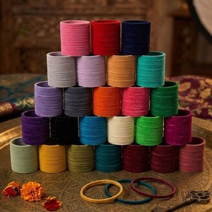 May include: A pyramid of colorful fabric bangles in shades of pink, red, blue, green, and more. The bangles are stacked on a decorative brass tray, with a few gold and teal bangles and a key in the foreground.