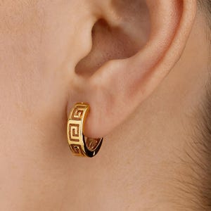Puede incluir: Un pendiente de aro dorado con un diseño de clave griega. El pendiente se lleva en la oreja y tiene un acabado brillante. El diseño es un patrón repetitivo de cuadrados y rectángulos.