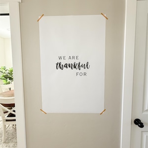 May include: A white rectangular sign with the words "WE ARE thankful FOR" in black script. The sign is attached to a beige wall with four small wooden clips. The background includes a doorway and a glimpse of a dining area.