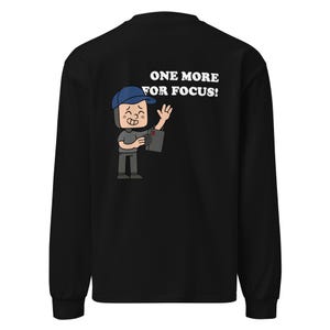 May include: Black long-sleeve shirt with a cartoon graphic on the back. The graphic features a character wearing a blue cap and holding a camera, with the text "ONE MORE FOR FOCUS!" in white.