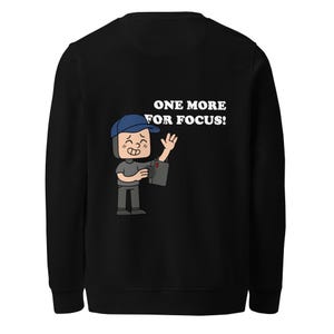 Może przedstawiać: Czarna bluza z kreskówkową ilustracją osoby trzymającej aparat fotograficzny. Osoba ta ma na sobie niebieską czapkę i szare ubrania. Tekst "ONE MORE FOR FOCUS!" jest wydrukowany na biało nad ilustracją.