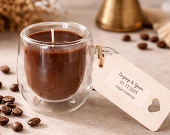 May include: A clear glass mug candle filled with brown wax, a white wick, and a small tag. The tag has text that reads "Zeynep & Yasin 01.12.2025 nişan hatırası" and a heart cutout. Coffee beans are scattered around the candle.