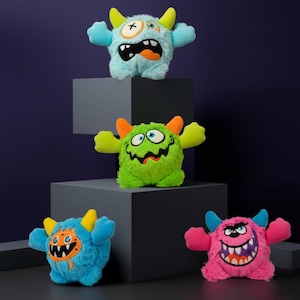 Puede incluir: Cuatro juguetes de peluche con forma de monstruo, con cuernos, brazos y caras expresivas, se exhiben sobre bloques geométricos grises sobre un fondo morado oscuro. Los monstruos son azules, verdes y rosas, con detalles en naranja y amarillo.