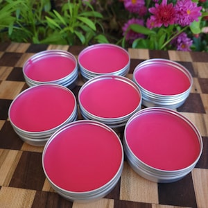 May include: Seven small, round, silver-colored tins filled with bright pink lip balm are arranged on a wooden surface. The lip balms have a smooth, glossy appearance. The background includes green foliage and pink flowers.
