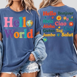 May include: A blue short-sleeve t-shirt featuring the text Hello World on the front with a globe graphic. The back displays various international greetings including Hello, Bonjour, Hola, Ciao, Aloha, Guten, Tag, Jambo, and Salut with flower icons.