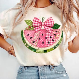 May include: A cream-colored t-shirt featuring a large graphic of a watermelon slice. The watermelon is decorated with pink and white gingham, floral, and heart patterns, topped with a pink gingham bow and green leaves with white polka dots.