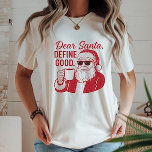 May include: Cream-colored t-shirt with a red graphic of Santa Claus wearing sunglasses and holding a mug. The text "Dear Santa, DEFINE GOOD." is above the Santa illustration. The shirt has short sleeves.
