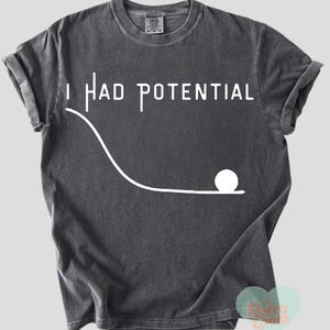 Puede incluir: Camiseta gris oscuro con el texto blanco "I HAD POTENTIAL" sobre un gráfico blanco de una bola rodando por un camino curvo. La camiseta tiene las mangas enrolladas y cuello redondo.