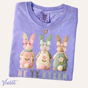 May include: Violet-colored t-shirt featuring three bunny figures with floral patterns and bows. The text "HE IS RYSEN" is printed below the bunnies. A silver necklace with a pendant is also visible.