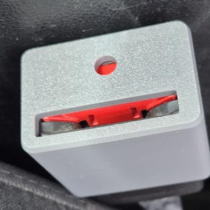 May include: A gray car seat belt buckle with a red release mechanism. The buckle is rectangular with a textured surface and a small hole at the top. The release mechanism is visible in the opening.