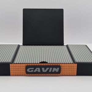 May include: A black and orange display stand with a gray surface made of small blocks. The stand has the name "GAVIN" in gray letters on the front. A black rectangular piece is attached to the back of the stand.