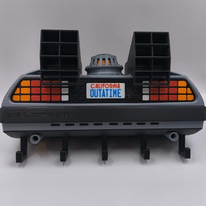 May include: A gray and black DeLorean-themed wall-mounted key holder. The key holder features the text "CALIFORNIA OUTATIME" on a license plate, with orange and red lights. It has four black hooks for keys.